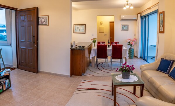 Ein Wohnzimmer mit einer Couch, einem Couchtisch mit Blumen, einem Essbereich mit Tisch und Stühlen sowie einer Küchenzeile mit Arbeitsfläche und Spüle.