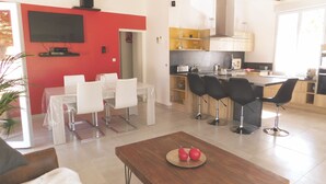 A modern living room with a wooden coffee table, a dining area with white chairs, and a kitchen with a red accent wall and wooden cabinets. - The oustaou of Sandrine (Valréas)