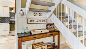 Un escalier agrémenté d’une console en bois et de tiroirs de rangement, un coin cuisine avec des meubles blancs et un plan de travail noir, et une enseigne indiquant « AMELIA ISLAND ».