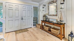 Un couloir avec un plancher en bois, une porte blanche, une console surmontée de paniers, un miroir avec un cadre en forme de coquillage et une enseigne indiquant « PLAGE ».
