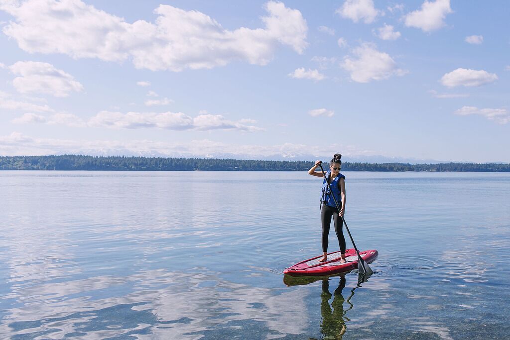 Une personne se tient debout sur une planche à pagaie dans une eau calme, avec une côte boisée en arrière-plan.