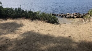 Blick auf einen See mit Sandstrand, felsigem Ufer und einem bewaldeten Hügel im Hintergrund.