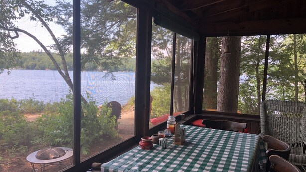 Ein Tisch mit kariertem Tuch, ein Korbstuhl und Blick auf einen See durch große Fenster.