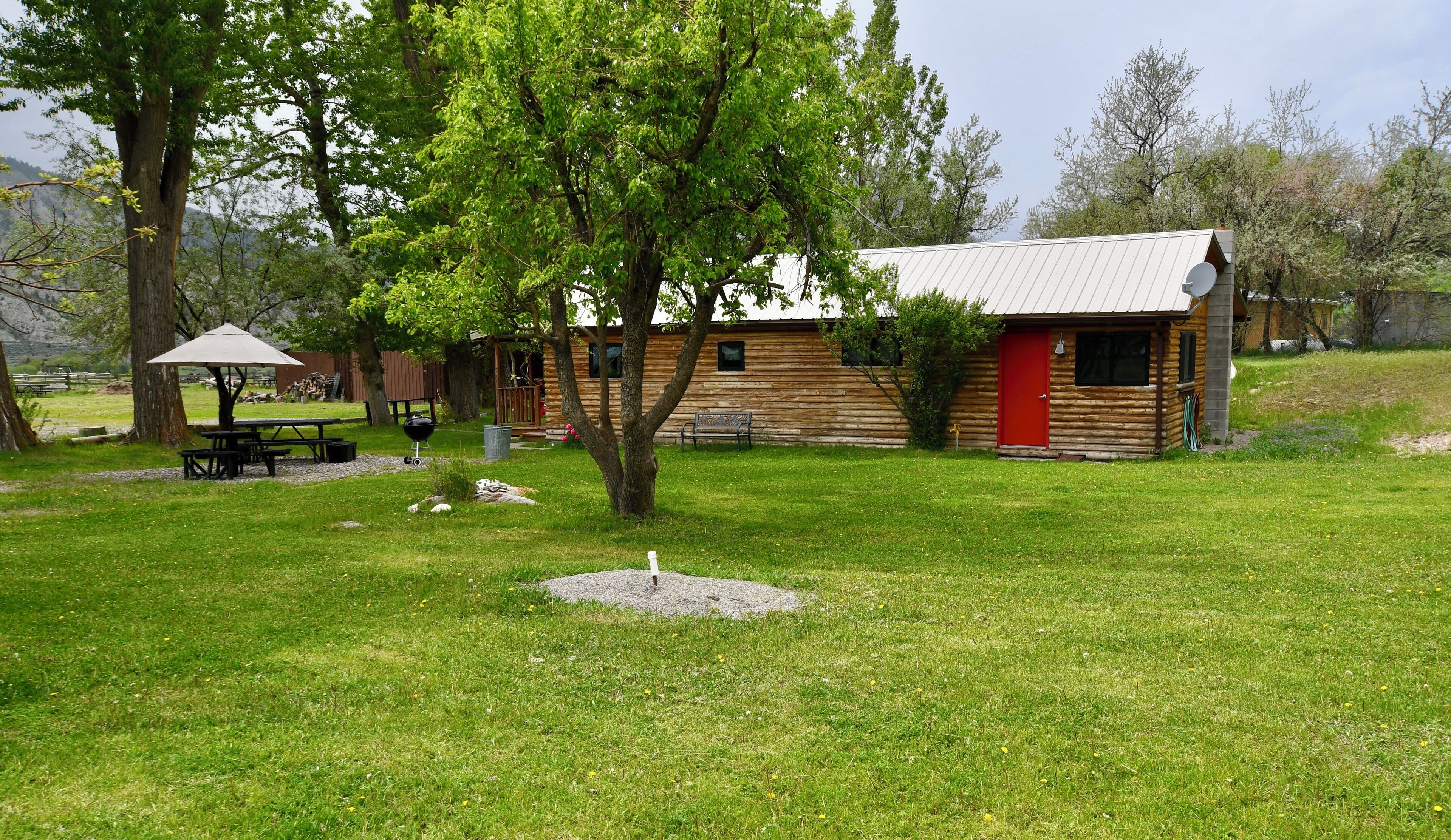 A rustic log cabin with a red door, surrounded by a grassy yard and trees.