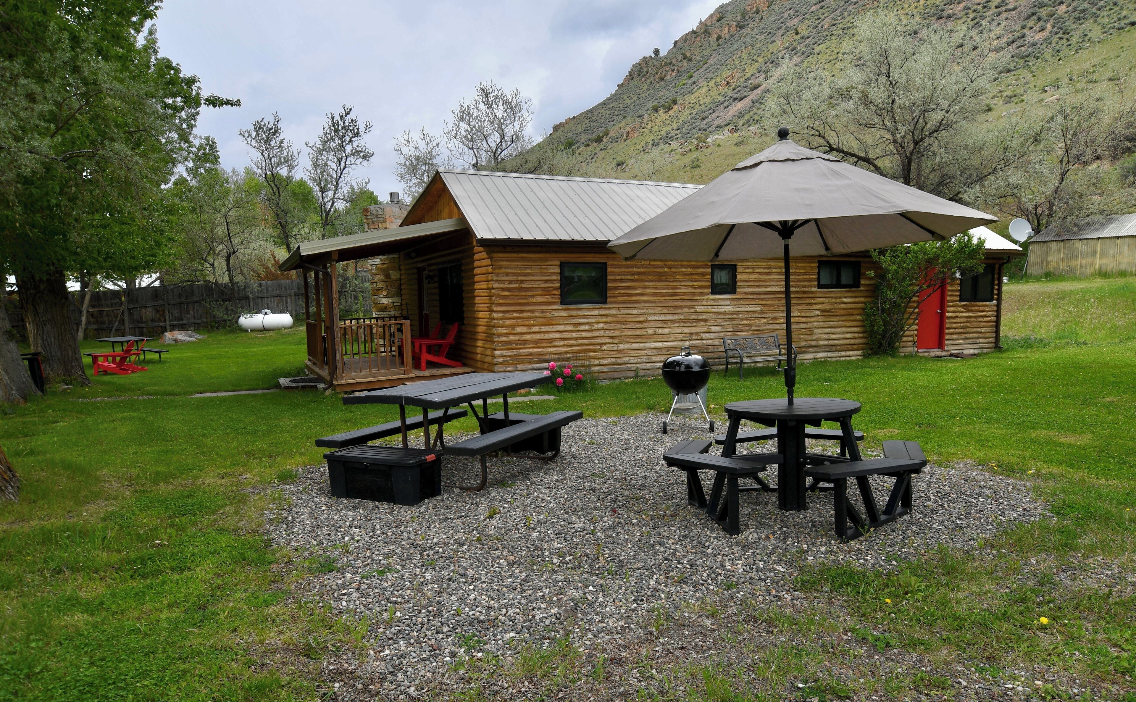 Une cabane rustique en rondins de bois, dotée d’un porche, de tables de pique-nique et d’un barbecue, située dans une cour arrière verdoyante.