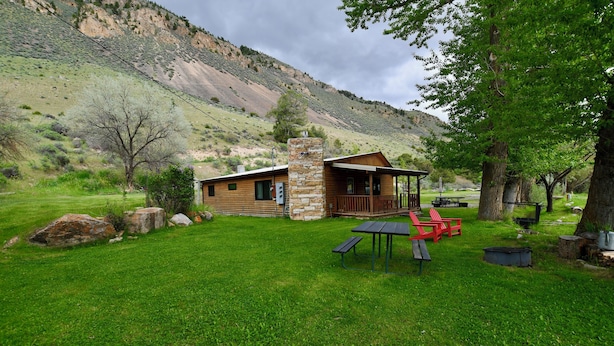 A rustic cabin with a stone chimney, surrounded by a grassy area with picnic tables and red chairs. - Rockin Y, Romantic Getaway or Family Friendly Lodging (Gardiner)