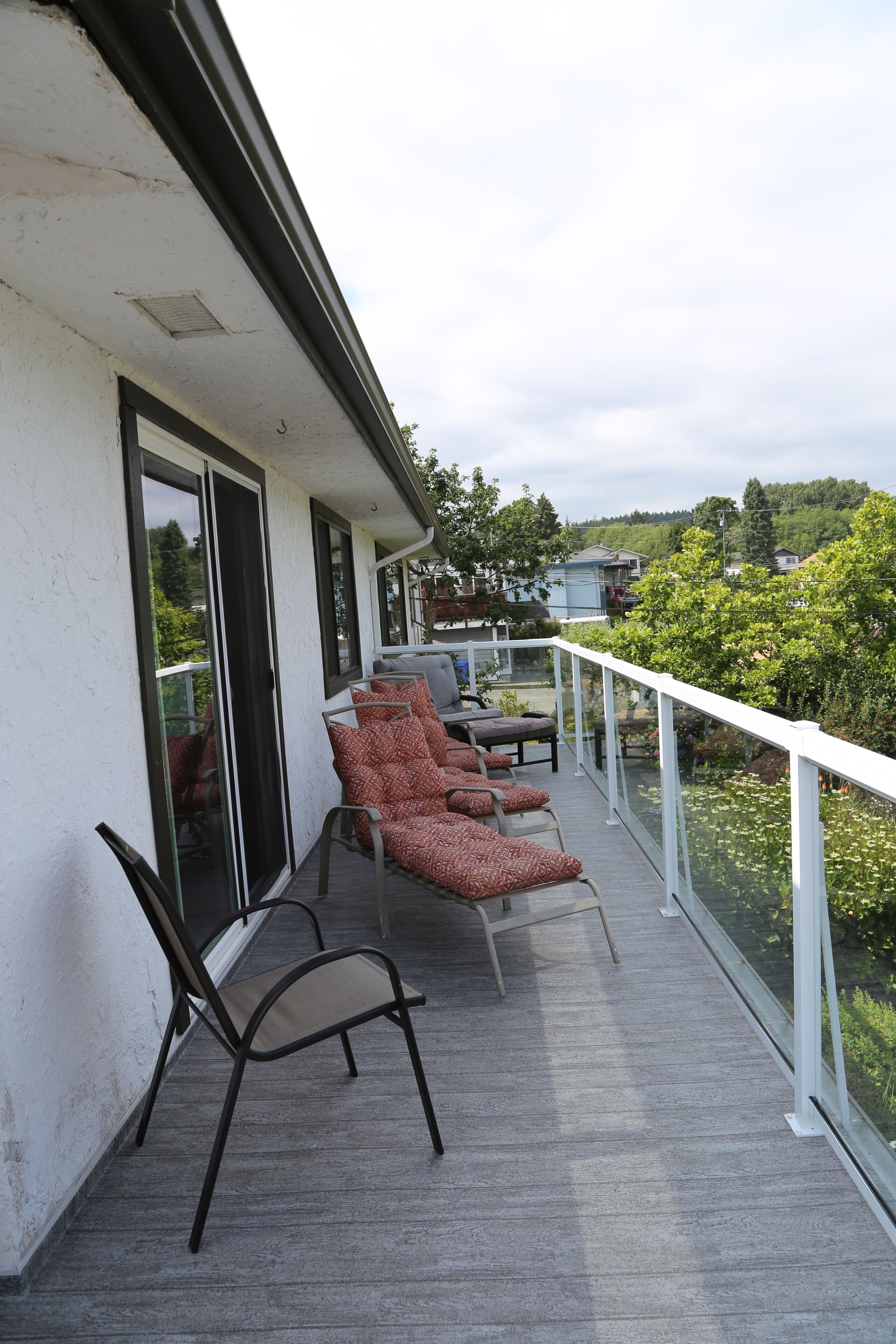 Un balcon avec une rangée de chaises et une table, donnant sur un jardin et un quartier résidentiel.