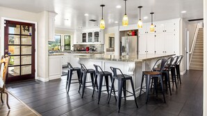 A modern kitchen with a central island, bar stools, pendant lighting, and a view of the outdoors. - Wine Country Estate with Vineyard Views, Pool, Spa, Fire-pit, 4 bedroom Suites (Temecula)