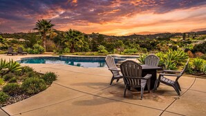 A pool area with chairs and a table, surrounded by greenery and a scenic view of hills and a sunset. - Wine Country Estate with Vineyard Views, Pool, Spa, Fire-pit, 4 bedroom Suites (Temecula)