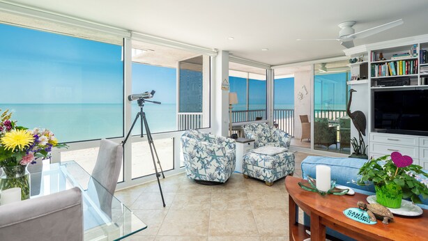 A spacious living area with a large glass door leading to a balcony overlooking the sea. There are two patterned armchairs, a glass coffee table, and a wooden side table with a candle and decorative items. - Ultimate Sanibel Beachfront! Island Beach Club, Top Floor, West-Facing, End Unit (Sanibel)