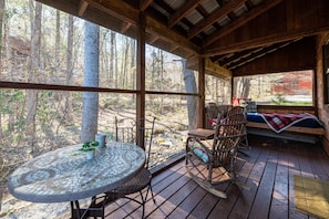 Relax in a serene outdoor space with a round table, wicker chairs, and a rocking chair, surrounded by wooden beams and forest views.