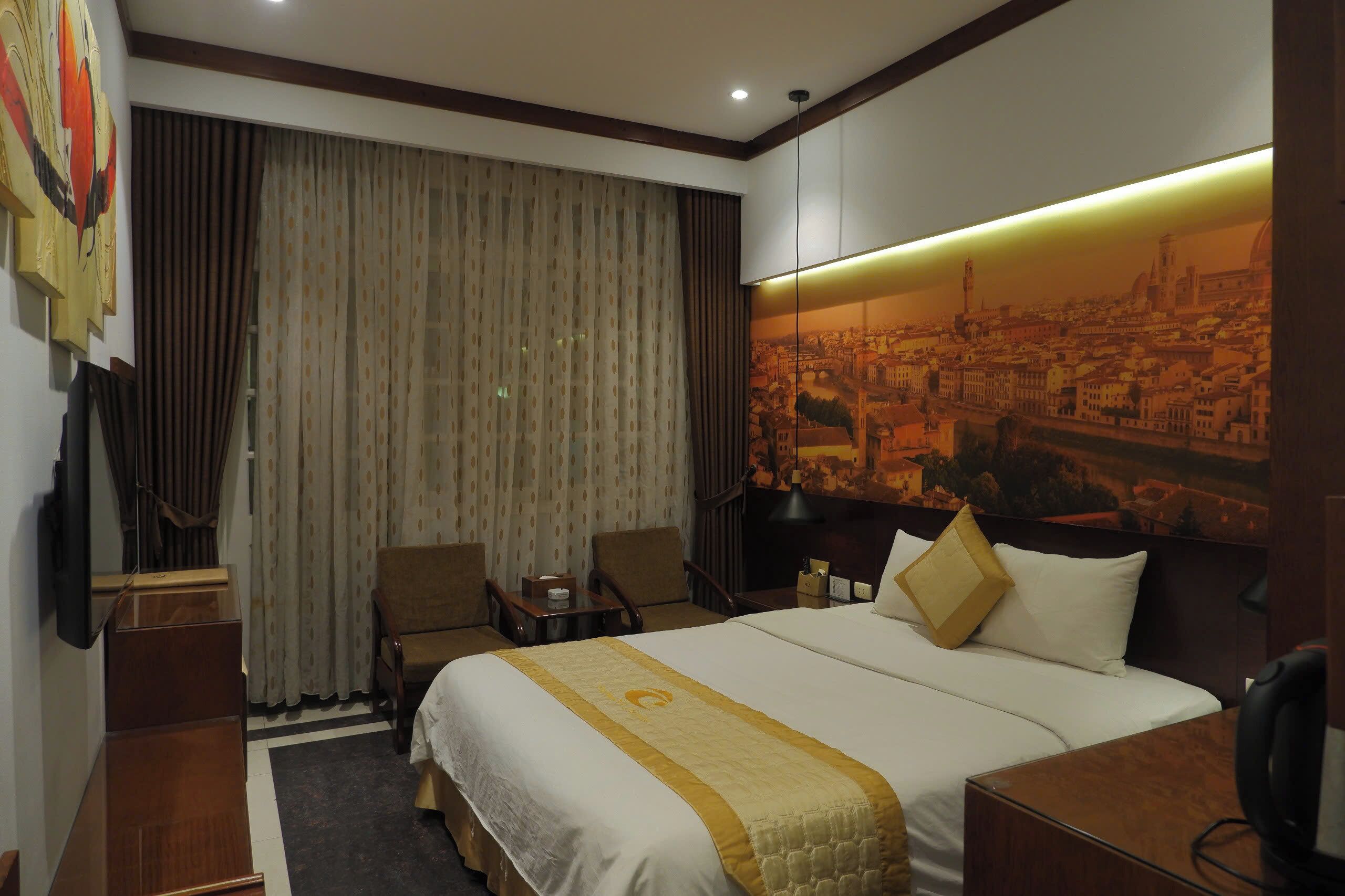 Habitación de hotel con una cama grande, un escritorio, una silla, un televisor y un mural de un paisaje urbano en la pared.