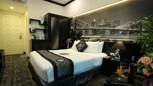 Une chambre d’hôtel avec un lit, une table de chevet, une lampe, une machine à café, un verre de jus d’orange, une bouteille d’eau et une fresque représentant un paysage urbain sur le mur.