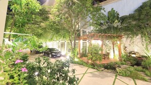 Un espace jardin bien éclairé, avec une voiture garée au fond, entouré d’une végétation luxuriante et agrémenté d’une pergola illuminée.