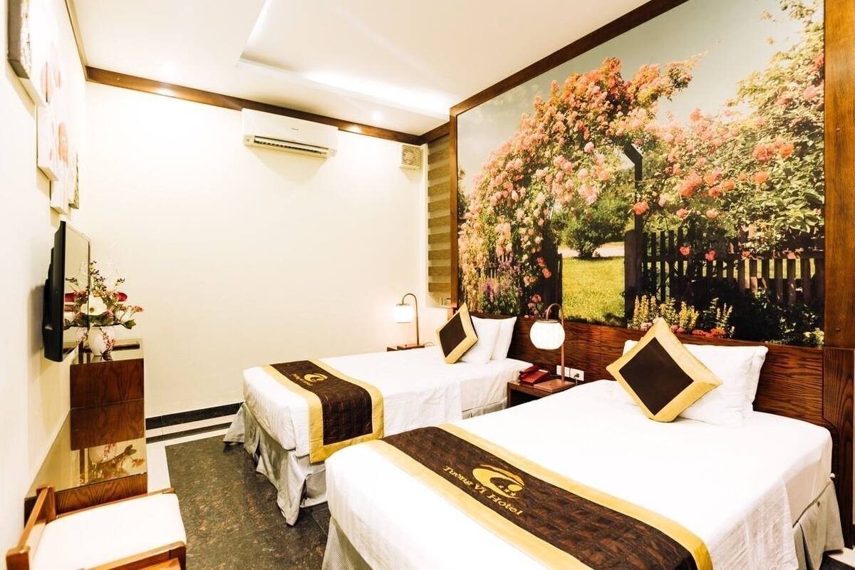 Une chambre d’hôtel avec deux lits, une grande fresque murale à motifs floraux, une tête de lit en bois, une table de chevet avec une lampe et un petit bureau avec une chaise.