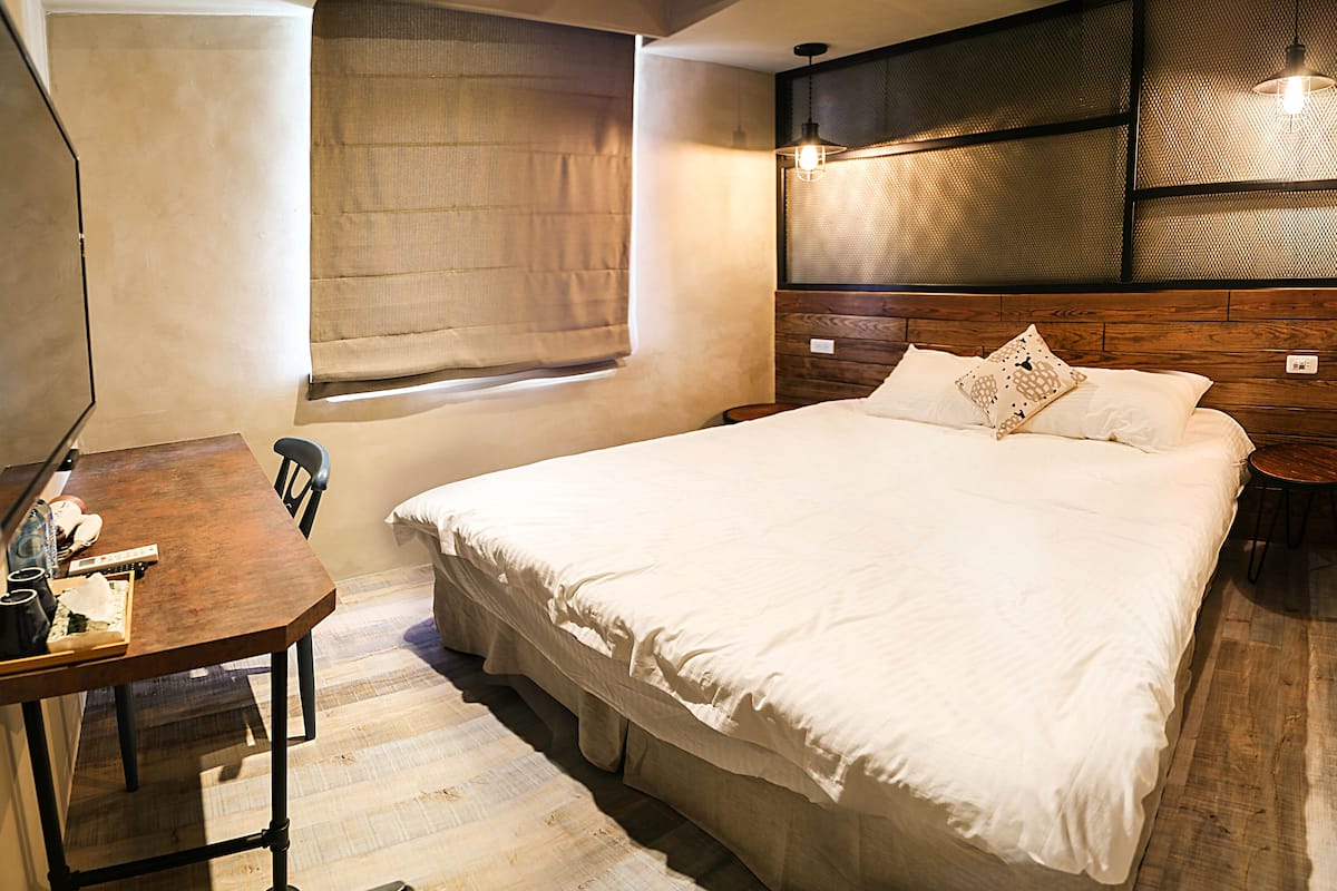 Un dormitorio con una cama grande, un escritorio de madera, una silla, una ventana con cortinas y una lámpara fijada a la pared.