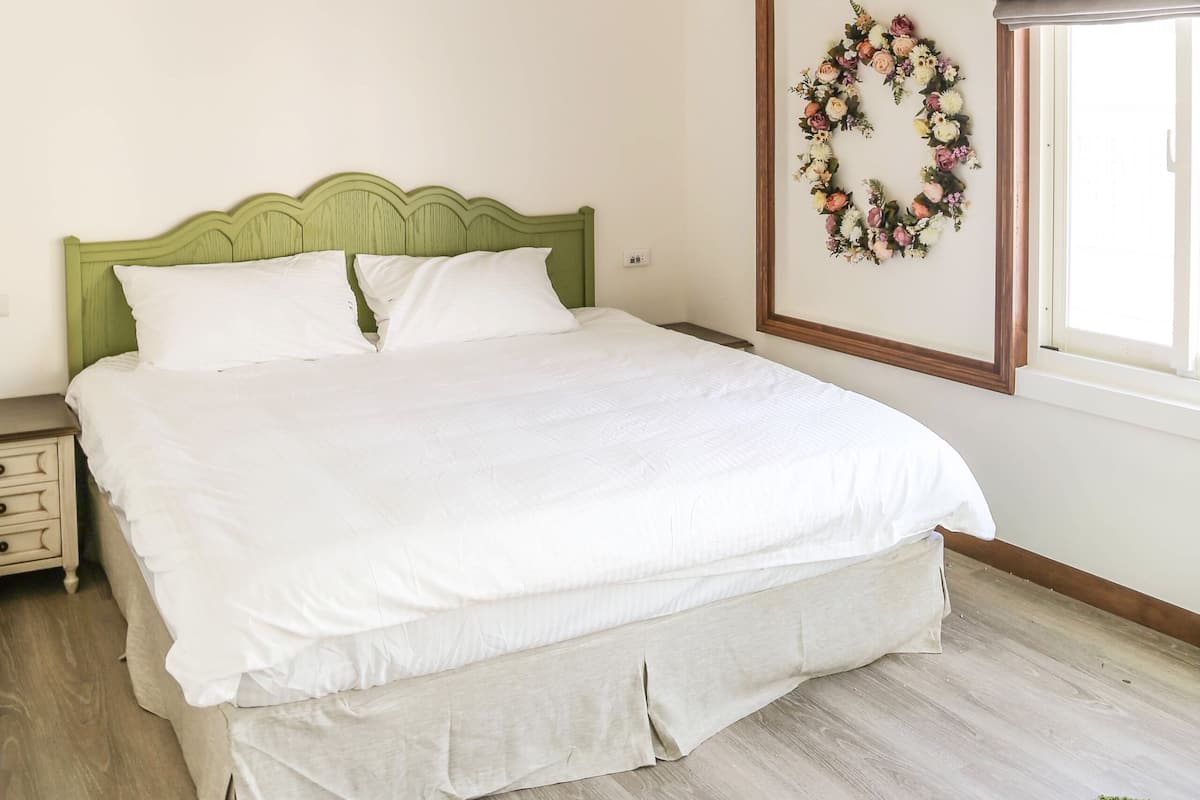 Un dormitorio con techo de madera, cabecera verde, cama blanca, una obra de arte floral enmarco y una ventana con vistas.