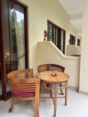 A balcony with a wooden table and chairs, a glass door, and a view of greenery. - ADI JAYA COTTAGES (Ubud)