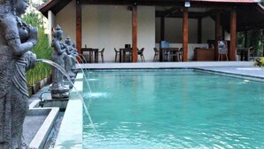 A pool with a statue on the left and a building with a red roof in the background. - ADI JAYA COTTAGES (Ubud)
