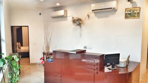 Un mostrador de recepción con una computadora, unidades de aire acondicionado y un ventilador. Hay una planta en maceta y una puerta que da a otra habitación.