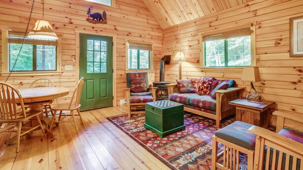A wooden cabin interior with a dining table, chairs, a sofa, and a small green ottoman.