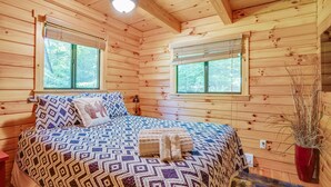 A wooden cabin bedroom with a patterned bedspread, two windows with blinds, a bedside lamp, and a potted plant.
