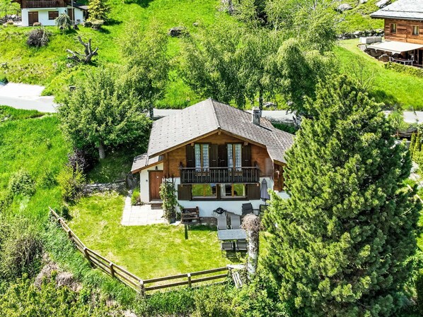 Une maison de style chalet dotée d’un balcon, entourée d’une végétation luxuriante et d’une clôture en bois.
