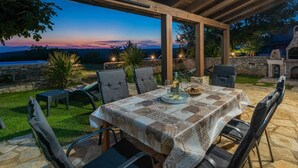 Een terras buiten met een eettafel, omringd door stoelen, onder een houten pergola.
