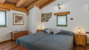Een slaapkamer met een houten plafond, een bed met een gewatteerde deken, twee nachtkastjes met lampen, een ingelijst schilderij aan de muur en een accentmuur van steen.