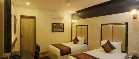 A hotel room with two single beds, a desk with a chair, a ceiling fan, an air conditioning unit, and a wall-mounted light fixture.