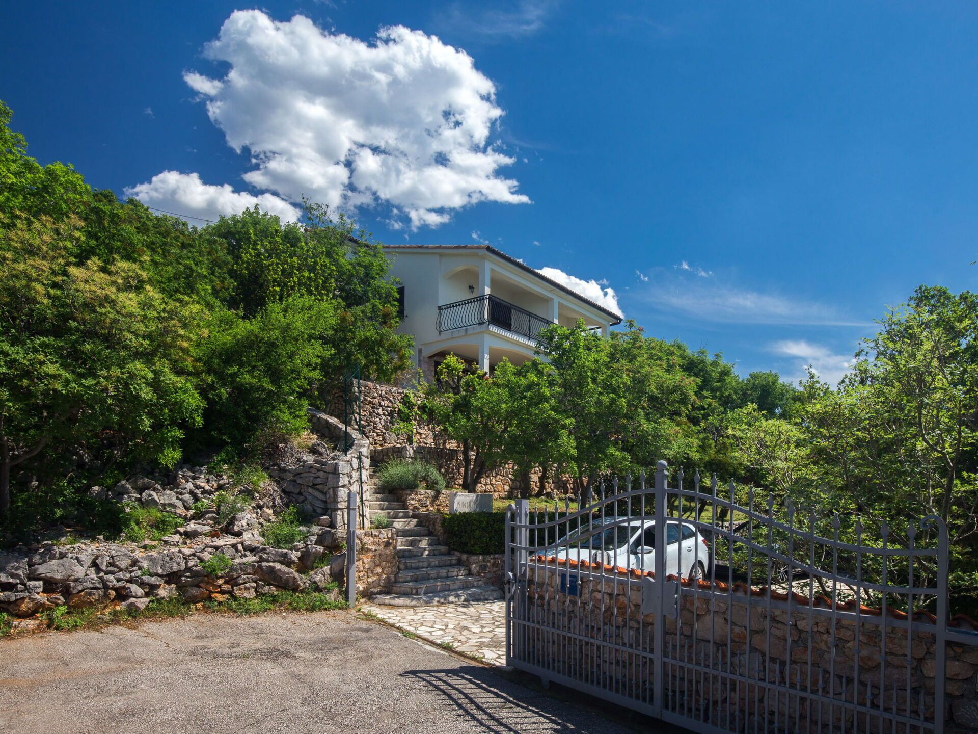 Ein zweistöckiges Haus mit einer Steintreppe zum Eingang, umgeben von üppiger Vegetation und blauem Himmel.