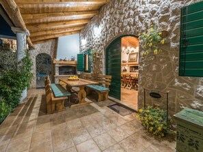 Un espace extérieur aménagé pour les repas, avec des murs en pierre, des bancs en bois et une table, une cheminée en pierre et une porte verte donnant sur l’intérieur.