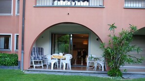 Un bâtiment doté d’un espace extérieur abrité avec des chaises en plastique blanches et une table, une petite plante et une entrée avec une porte en verre.