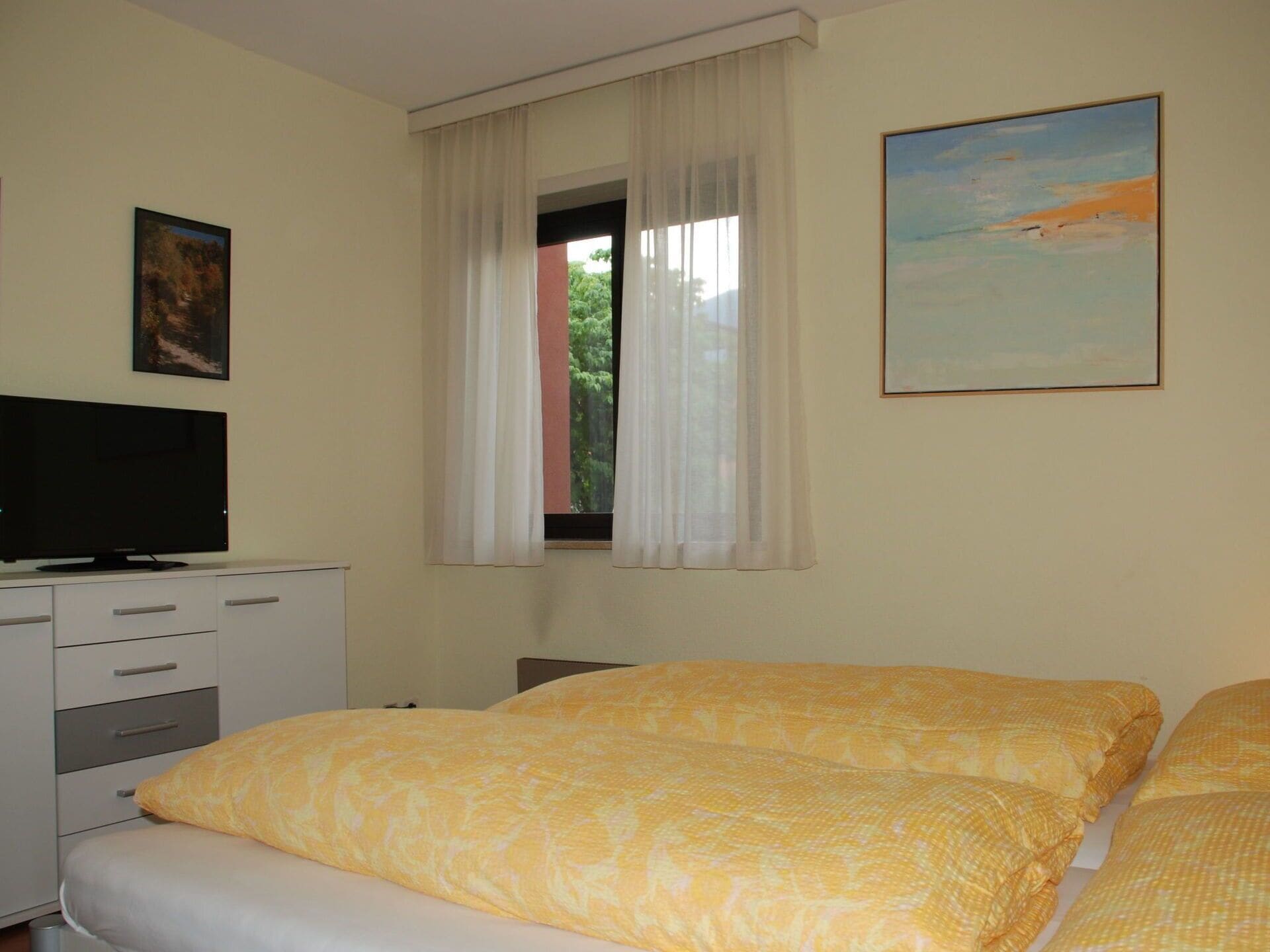 A bedroom with a bed, a TV, a window with curtains, and a painting on the wall.