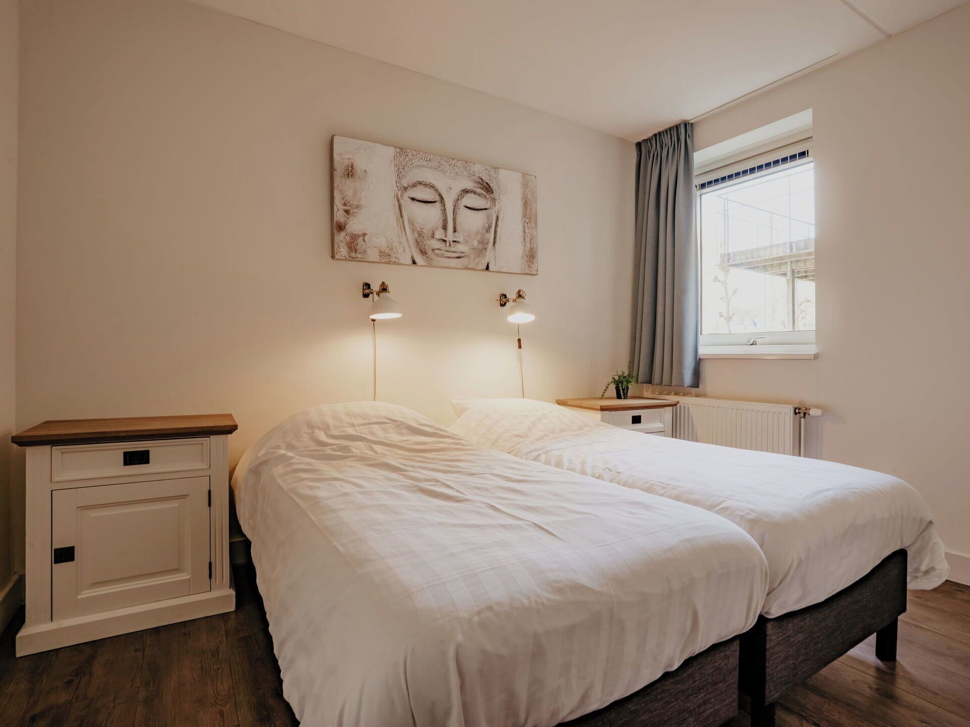 A bedroom with two beds, a nightstand, a wall-mounted lamp, a window with curtains, and a radiator.