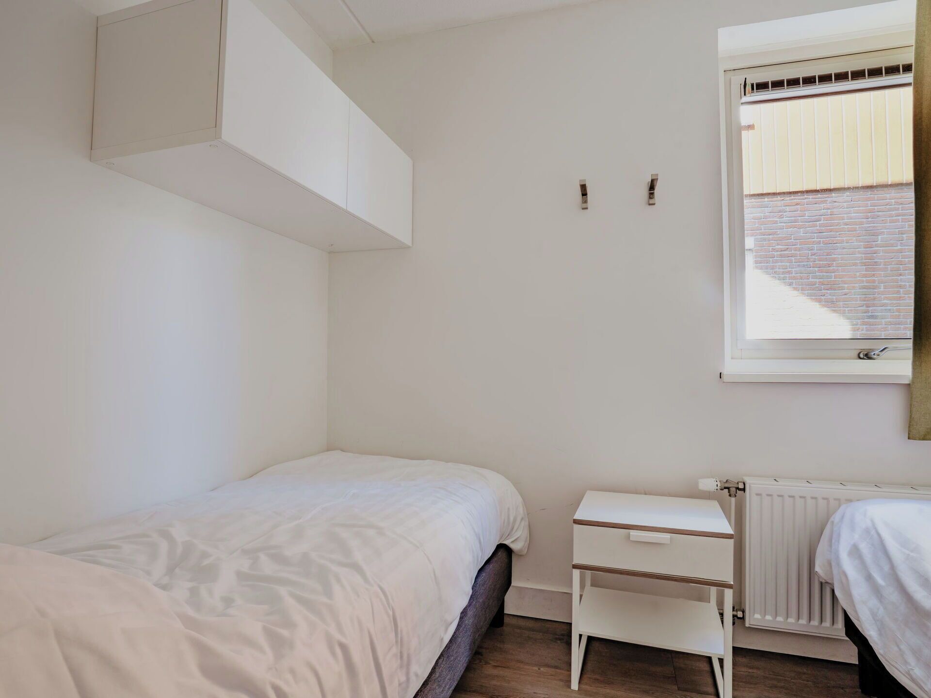 Een slaapkamer met een bed, een klein nachtkastje, een radiator en een raam met uitzicht op een bakstenen muur.