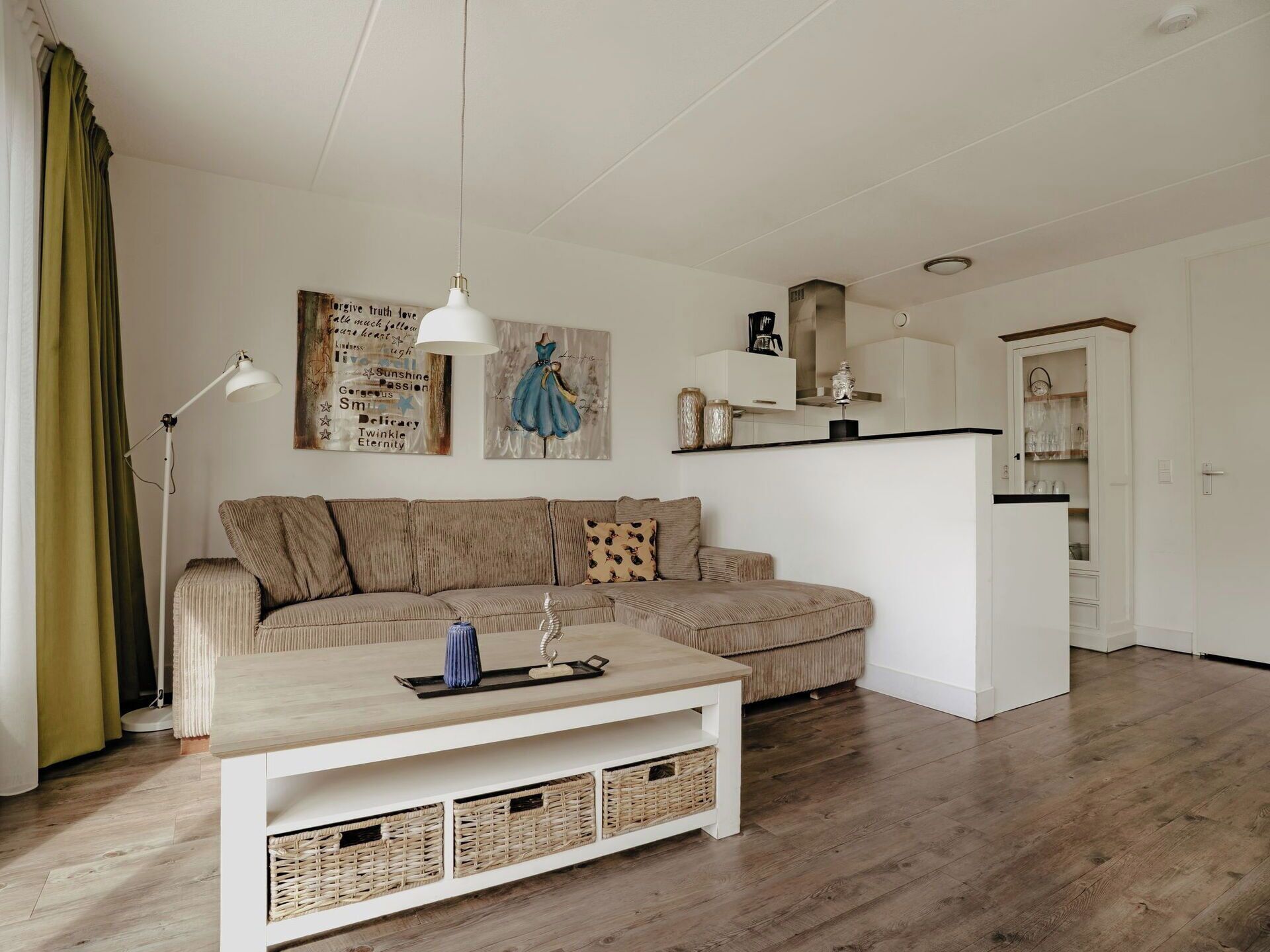 Een moderne woonkamer met een hoekbank, een salontafel, een vloerlamp en een keukenhoek met kasten en een koelkast.