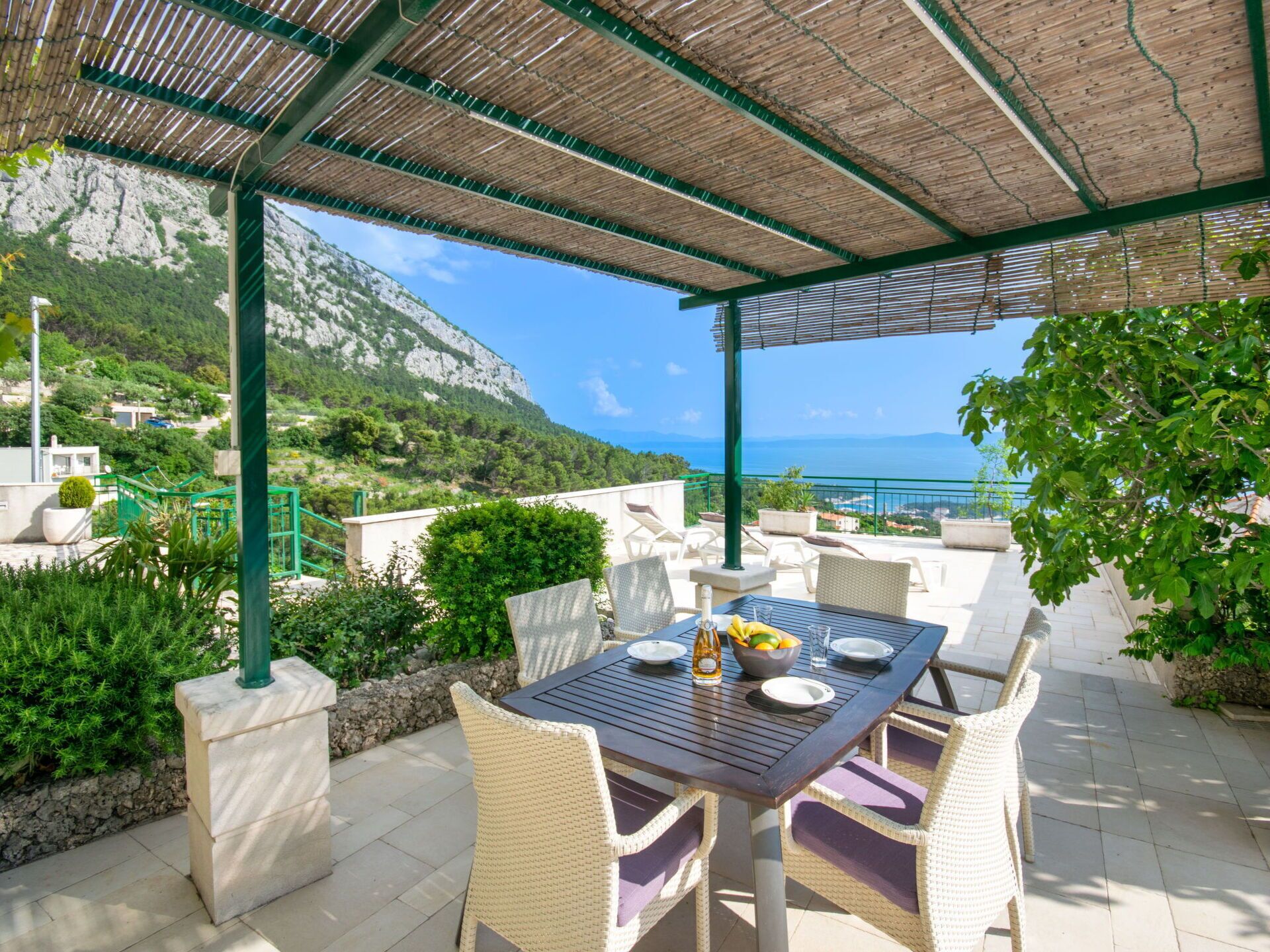 An outdoor patio with a table set for dining, surrounded by wicker chairs, overlooking a scenic coastal view with mountains in the background.