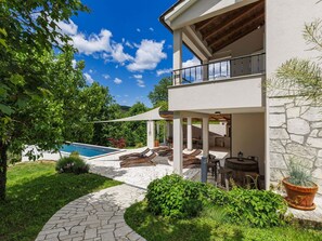 A modern two-story house with a stone exterior, a pool, outdoor seating, and lush greenery.