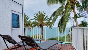 A wooden deck with two lounge chairs, a white door, and a view of the ocean with palm trees. - Boutique vllla on the rocks (Trou d'Eau Douce)