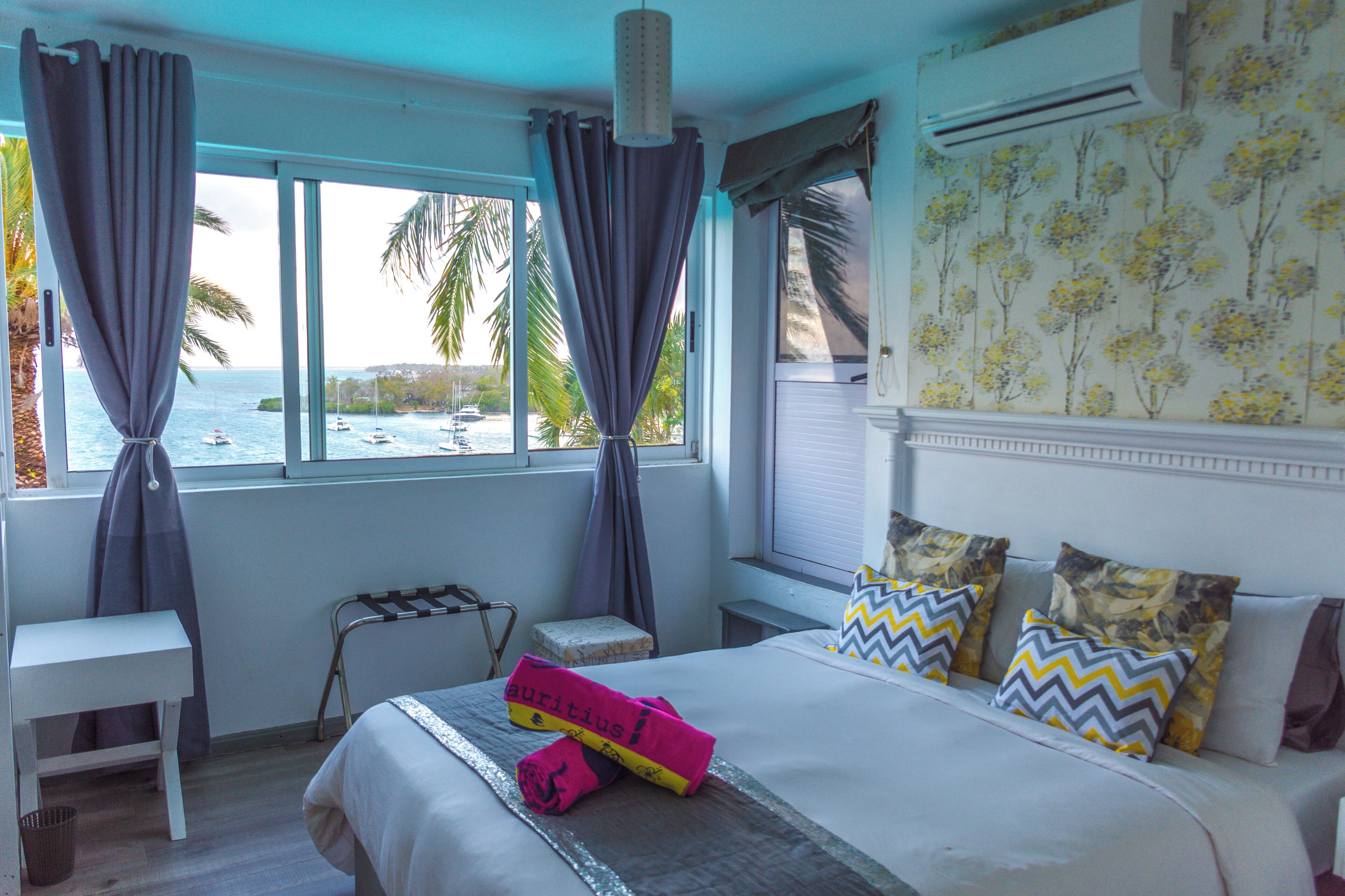 Habitación de hotel con cama, un banco, un taburete pequeño y vista al mar a través de amplias ventanas.