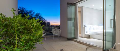 A modern hotel room with a glass door leading to a balcony, a bed, and a seating area with chairs.