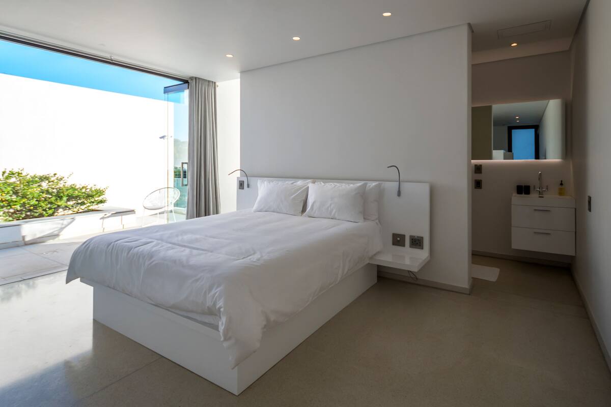 A modern bedroom with a large bed, a glass door leading to a balcony, and a minimalist bathroom.
