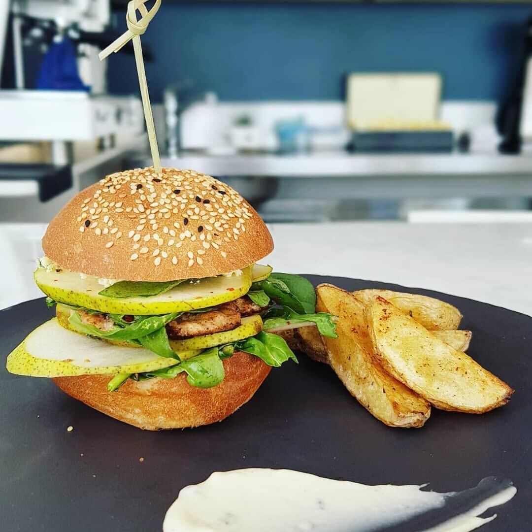 En burger med sesamfrøbolle, salat og syltede agurker, serveret med kartoffelbåde og en portion sauce.