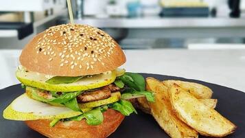 En burger med sesamfrøbolle, salat og syltede agurker, serveret med kartoffelbåde og en portion sauce.