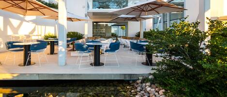 Un área de comedor al aire libre con mesas, sillas, un elemento acuático y follaje.