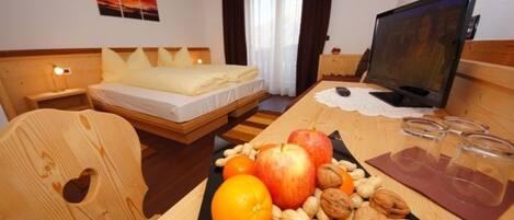 A hotel room with a bed, wooden headboard, a desk with a computer, a fruit and nut display, and a window with curtains.