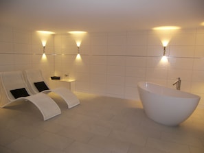 Une salle de bain moderne avec une baignoire sur pieds, deux fauteuils et des appliques murales.