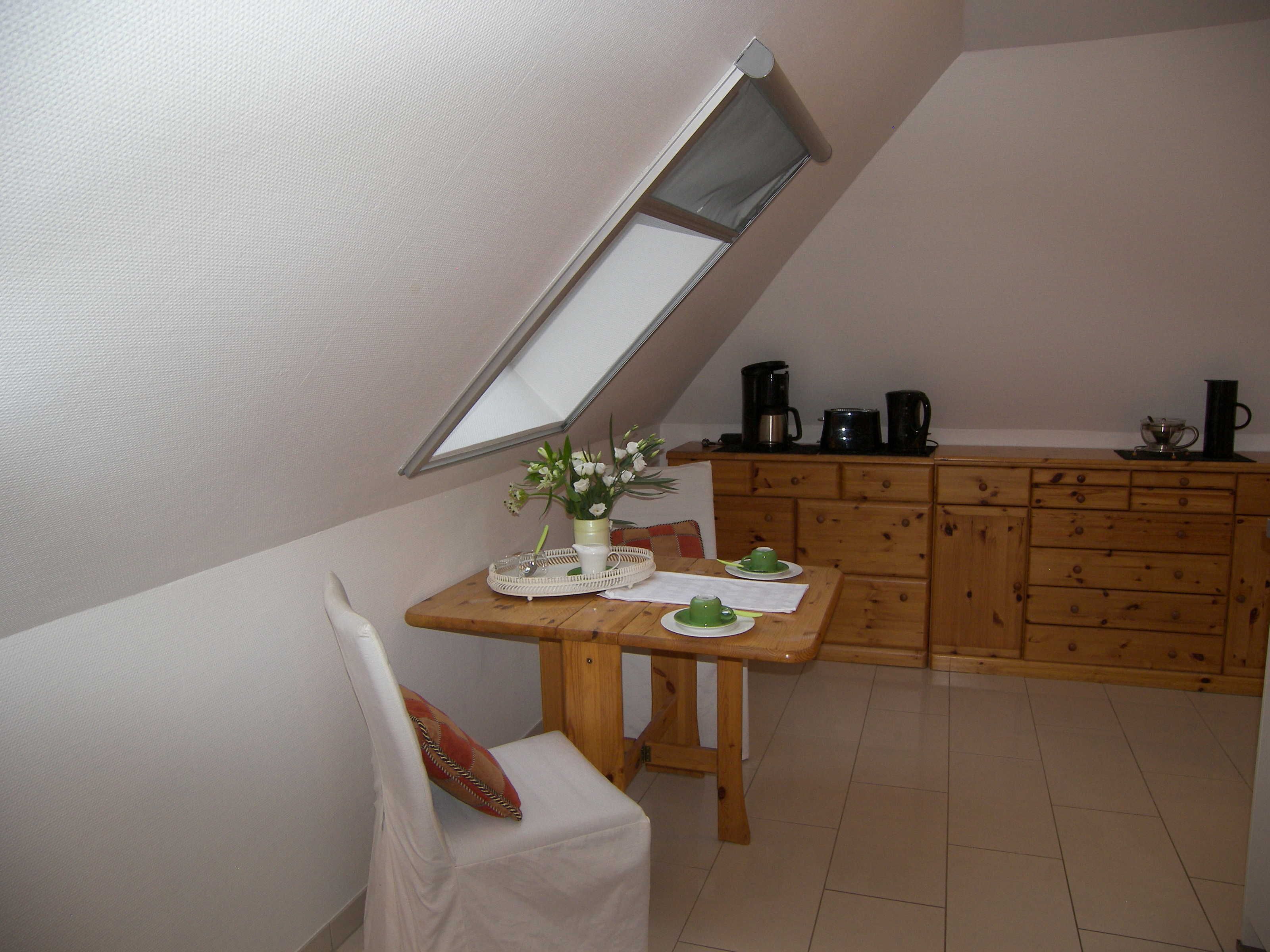 A small, cozy room with a slanted ceiling, a wooden dining table with a vase of flowers, a chair, and a kitchen area with cabinets and a coffee machine.