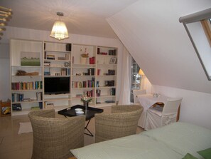 A cozy living room with a bookshelf, a television, wicker chairs, and a dining table.